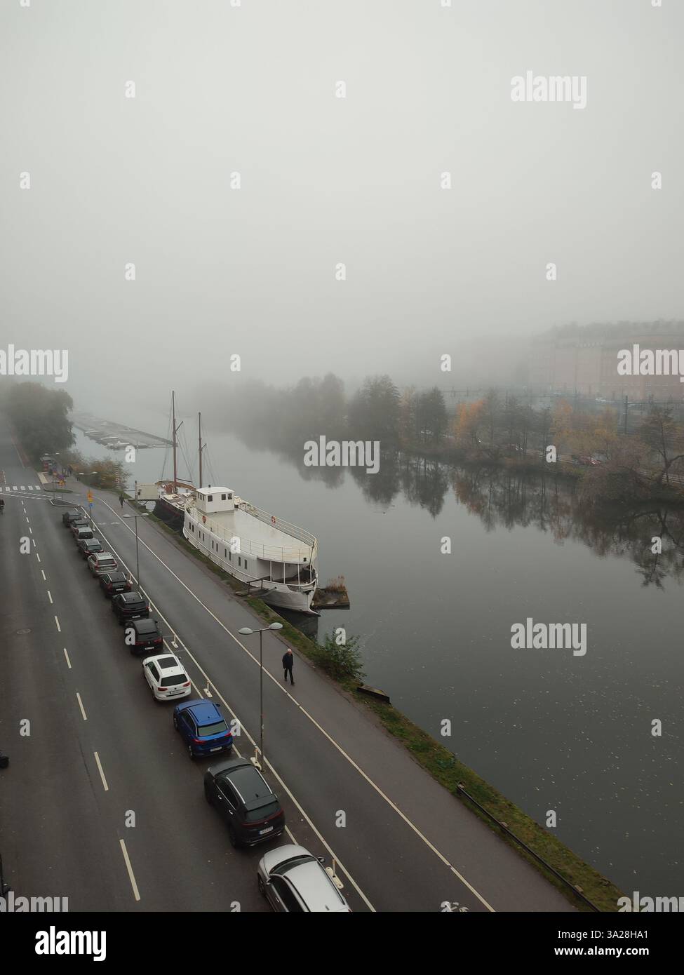 Nebeliger Morgenblick auf ein angedocktes Boot und ein baumgesäumtes Flussbett im Spätherbst im Zentrum von Stockholm, Schweden - Smartphone-aufgenommenes Stockfoto