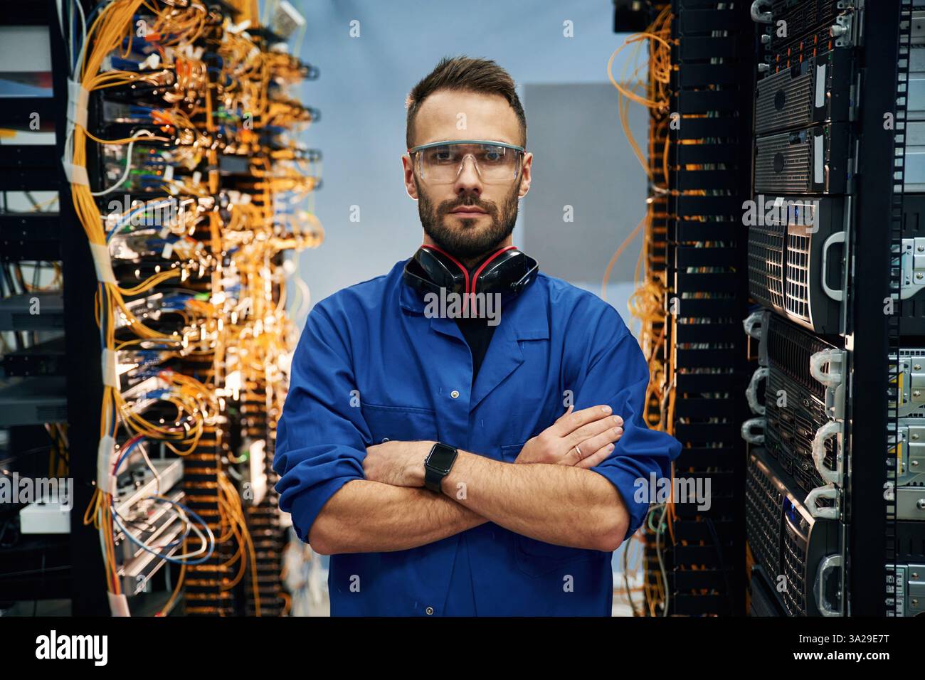 Stehend mit gekreuzten Armen. Der junge Mann arbeitet mit Internetgeräten und Kabeln im Serverraum. Stockfoto