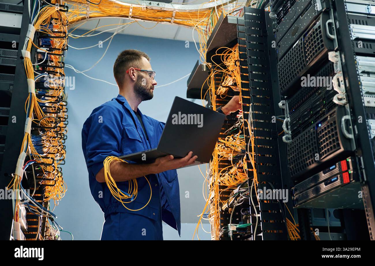 Mit Laptop in der Hand. Der junge Mann arbeitet mit Internetgeräten und Kabeln im Serverraum. Stockfoto