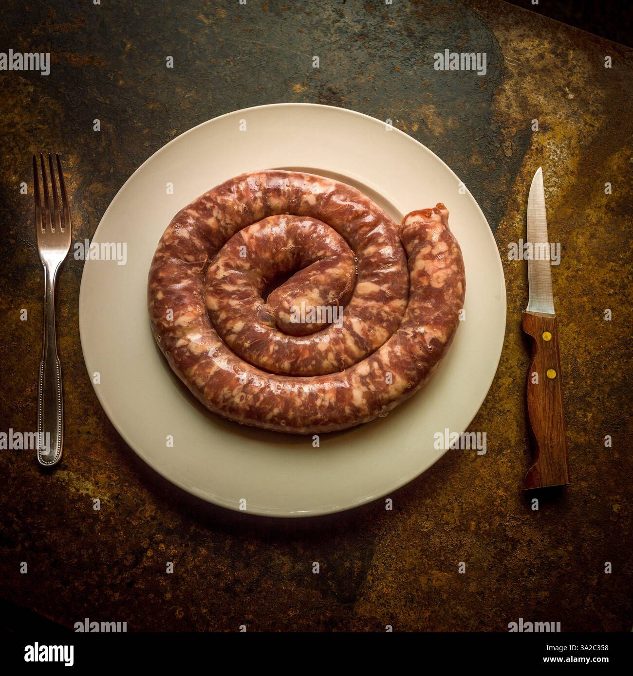 Schweinewurst auf einem weißen Teller, umgeben von einem Messer und einer Gabel an einem rustikalen Tisch Stockfoto