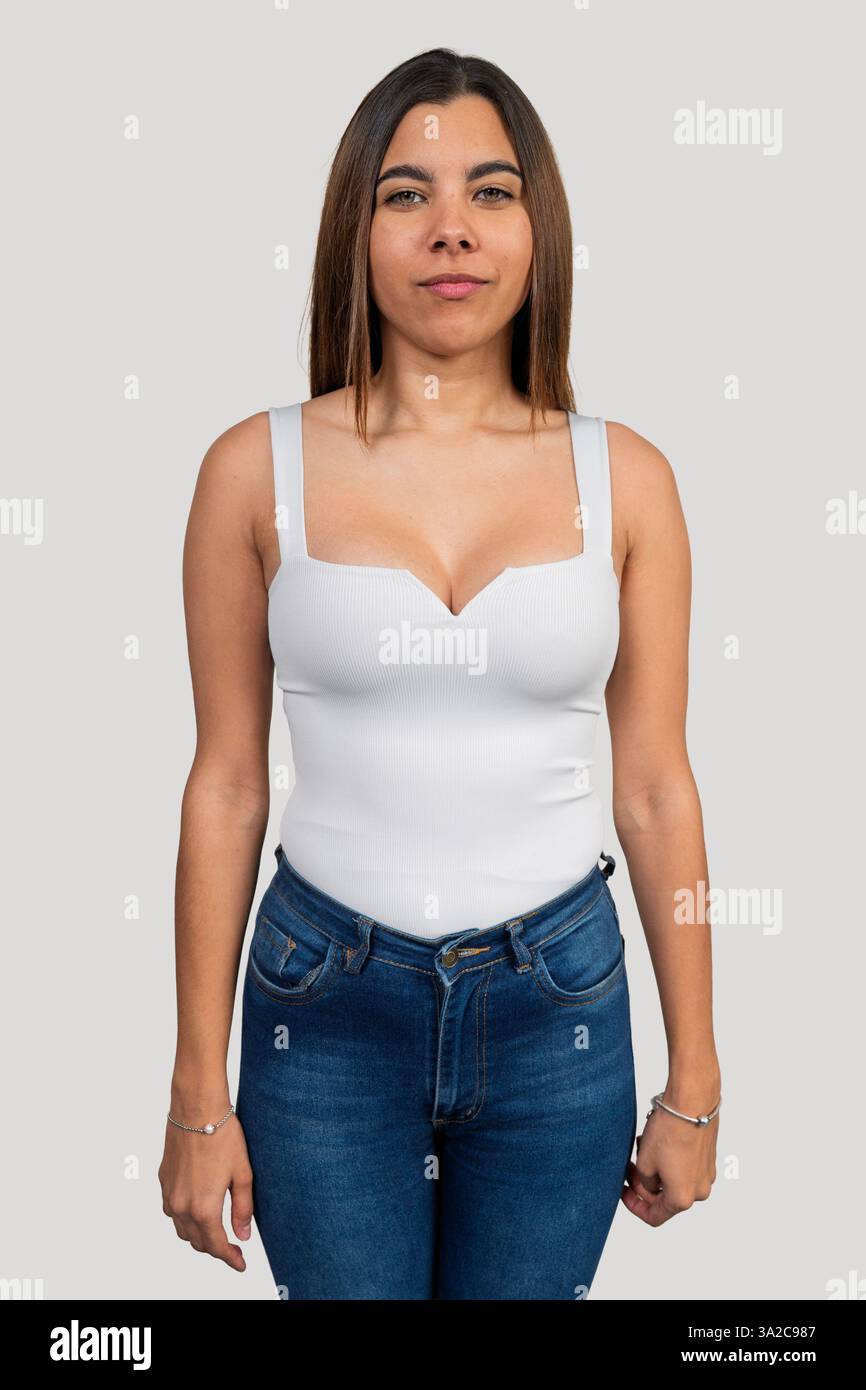 Studio-Porträt einer jungen lateinfrau mit weißem Oberteil und Jeans, die selbstbewusst vor einem schlichten Hintergrund steht Stockfoto