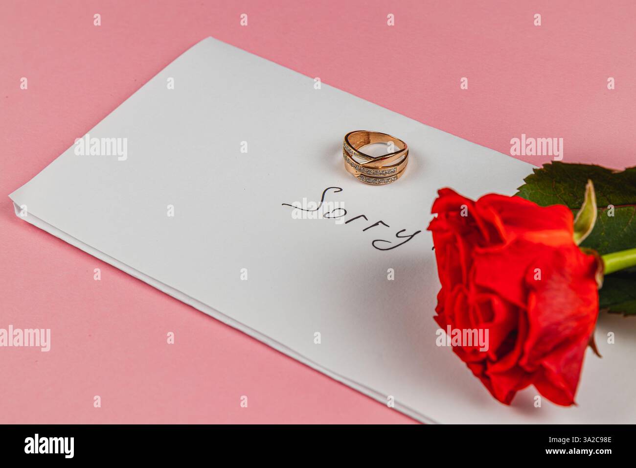 Ein goldener Verlobungsring sitzt auf einem weißen Umschlag, auf dem das Wort Sorry handgeschrieben ist. Neben dem Ring fügt eine rote Rose einen Hauch von Emotionen hinzu, A Stockfoto