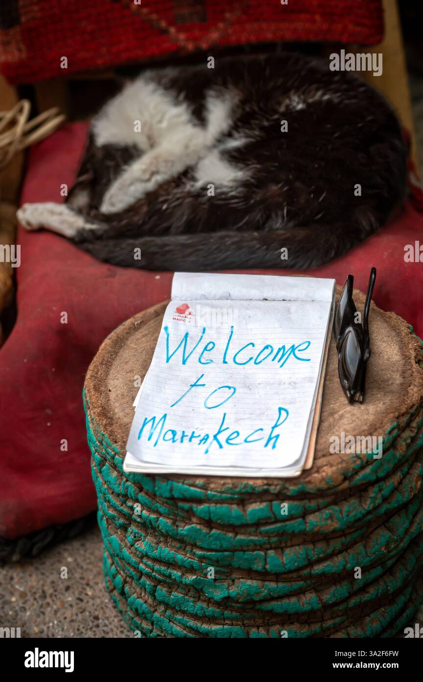 Ein gemütliches Willkommensschild liegt auf einem Tisch, während eine Katze in der Nähe von Marrakesch schläft. Stockfoto