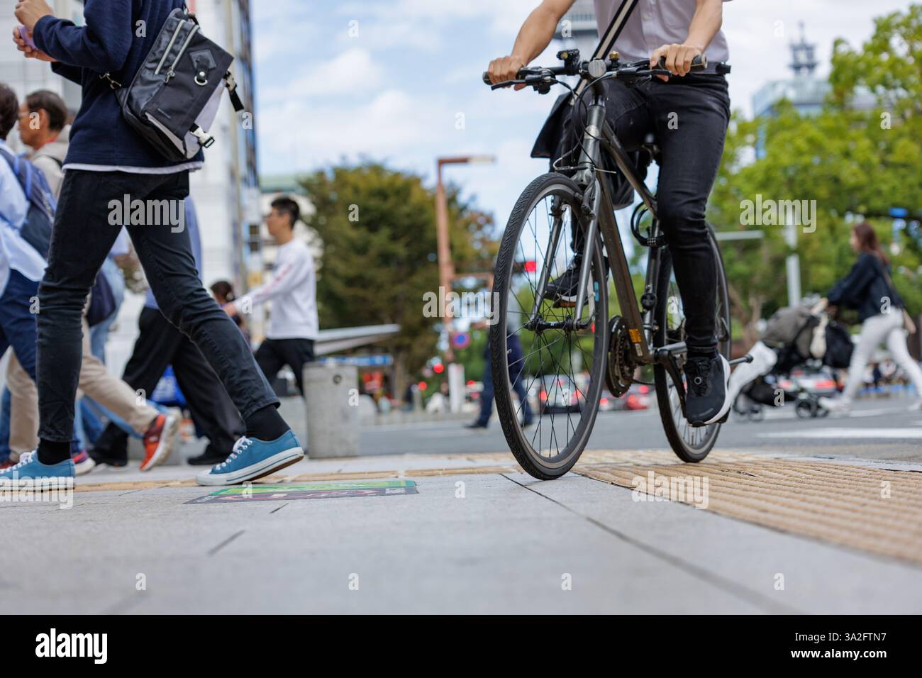Radfahrer fahren durch die Stadtüberquerung voller Fußgänger, die auf einem von Bäumen gesäumten Boulevard schlendern. Die Szene fängt die Hektik des Stadtlebens ein. Stockfoto