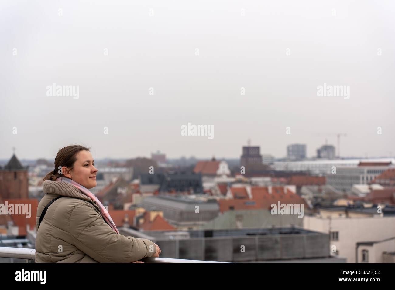 Lächelnde Frau in warmer Jacke und Schal, lehnt sich auf Geländer und genießt an einem bewölkten Tag einen Panoramablick auf die Stadt. Stockfoto