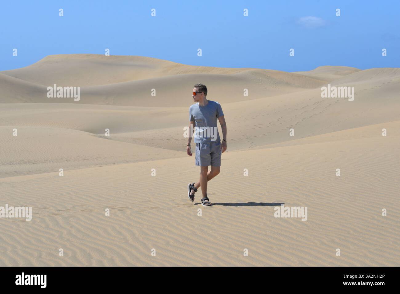 Ein junger Mann, der an einem heißen, sonnigen Tag auf einer Sanddüne durch Barhan in Maspalomas, Gran Canaria, spaziert. Touristisches Konzept für Wüstenreisen. Stockfoto