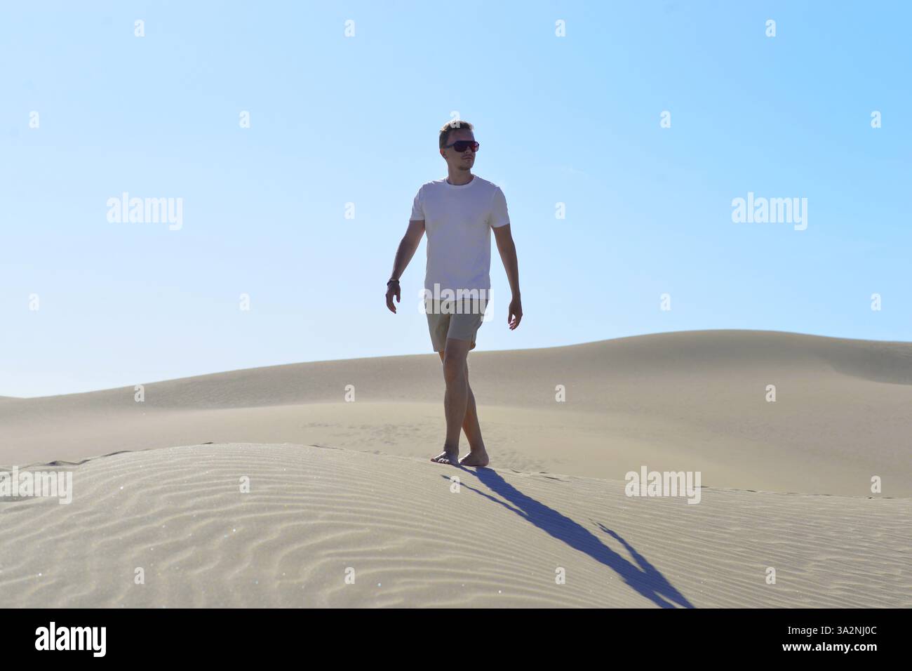 Ein junger Mann, der an einem heißen, sonnigen Tag auf einer Sanddüne durch Barhan in Maspalomas, Gran Canaria, spaziert. Touristisches Konzept für Wüstenreisen. Stockfoto