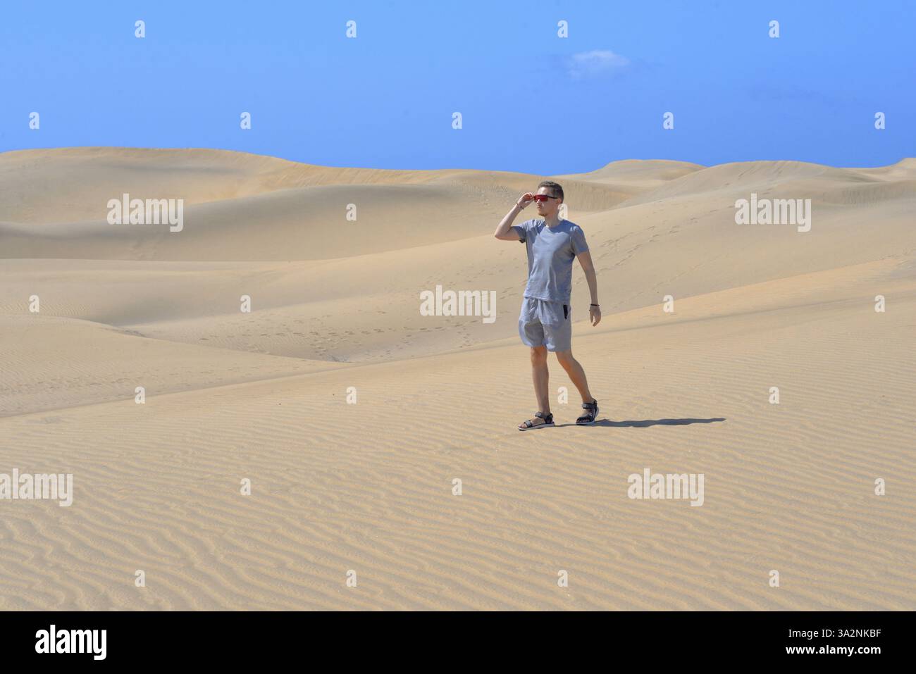Ein junger Mann, der an einem heißen, sonnigen Tag auf einer Sanddüne durch Barhan in Maspalomas, Gran Canaria, spaziert. Touristisches Konzept für Wüstenreisen. Stockfoto
