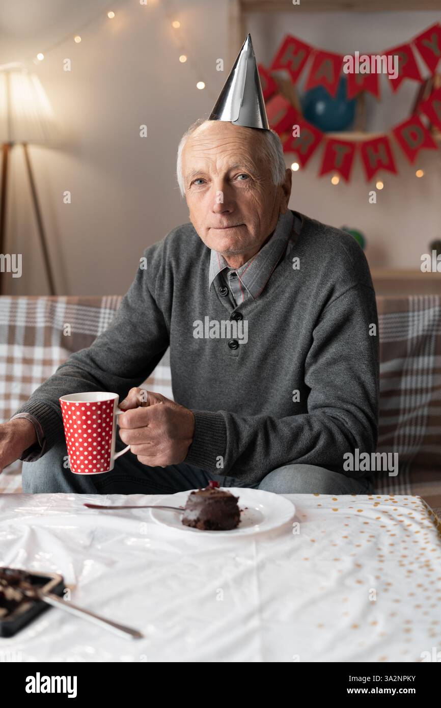 Porträt eines pensionierten Mannes mit Partyhut, der allein Geburtstag feiert und mit Traurigkeit in die Kamera schaut Stockfoto
