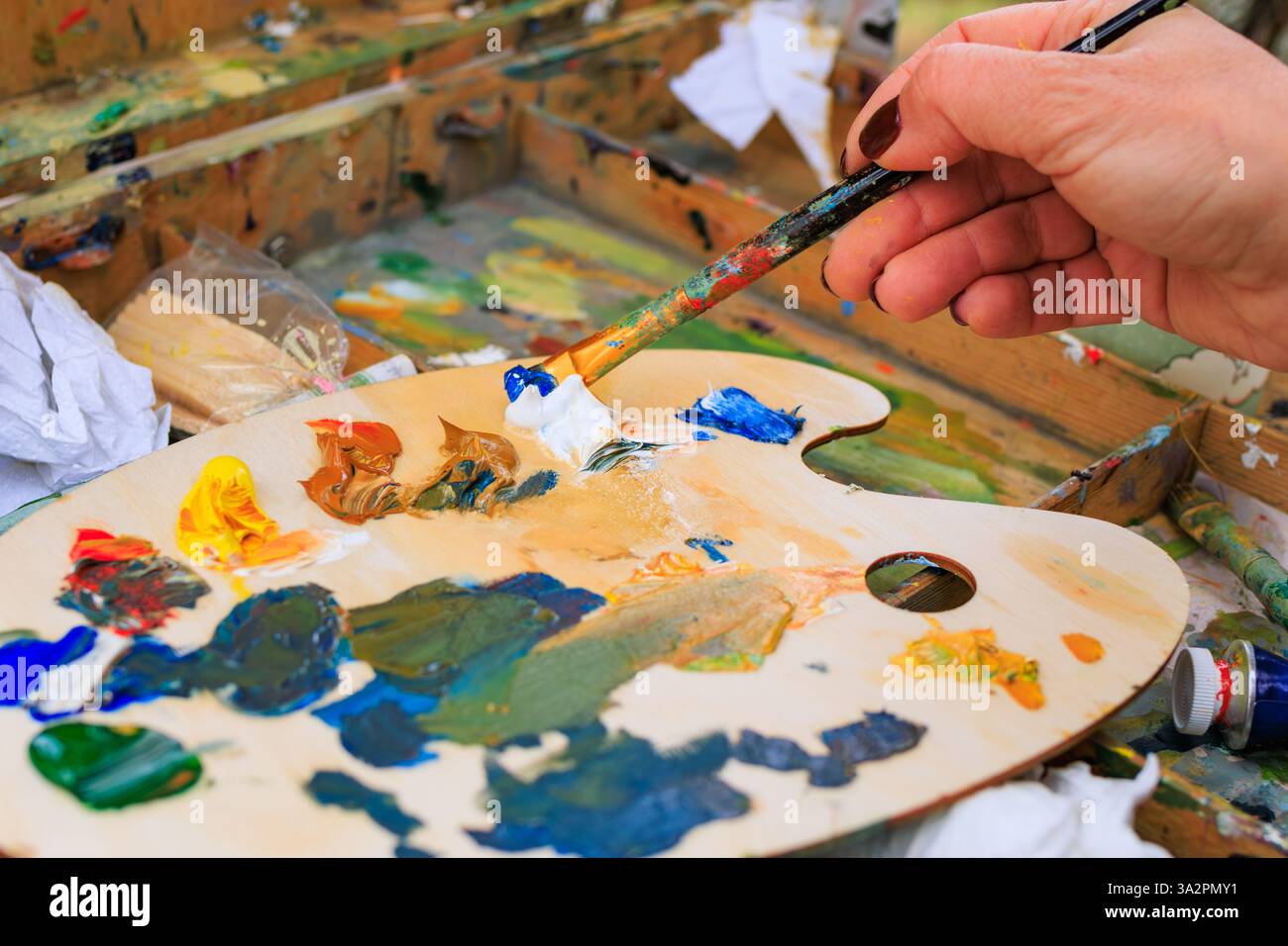 Ein Künstler mischt mit Hand blaue und weiße Farbe auf einer Holzpalette mit einem Pinsel, umgeben von lebhaften Farben und einem unordentlichen Arbeitsbereich, um die Kreationen festzuhalten Stockfoto