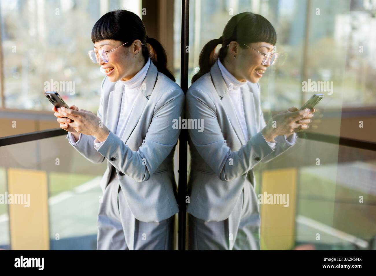 Eine fröhliche professionelle asiatische Frau in einem stilvollen grauen Anzug lächelt weit, während sie ihr Handy hält. Sie steht in einem modernen Büro mit großem Windo Stockfoto