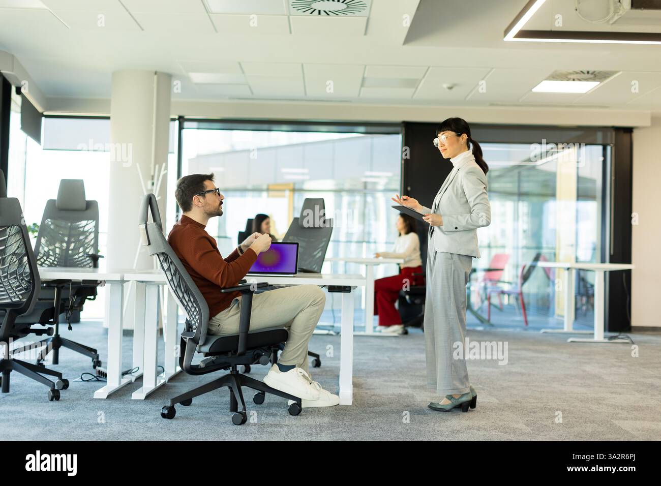 In einem modernen Büro arbeitet eine Frau in einem Anzug mit einem sitzenden Mann zusammen, während Kollegen an Schreibtischen in der Nähe arbeiten, um kreative Diskussionen und Teamarbeit zu fördern Stockfoto