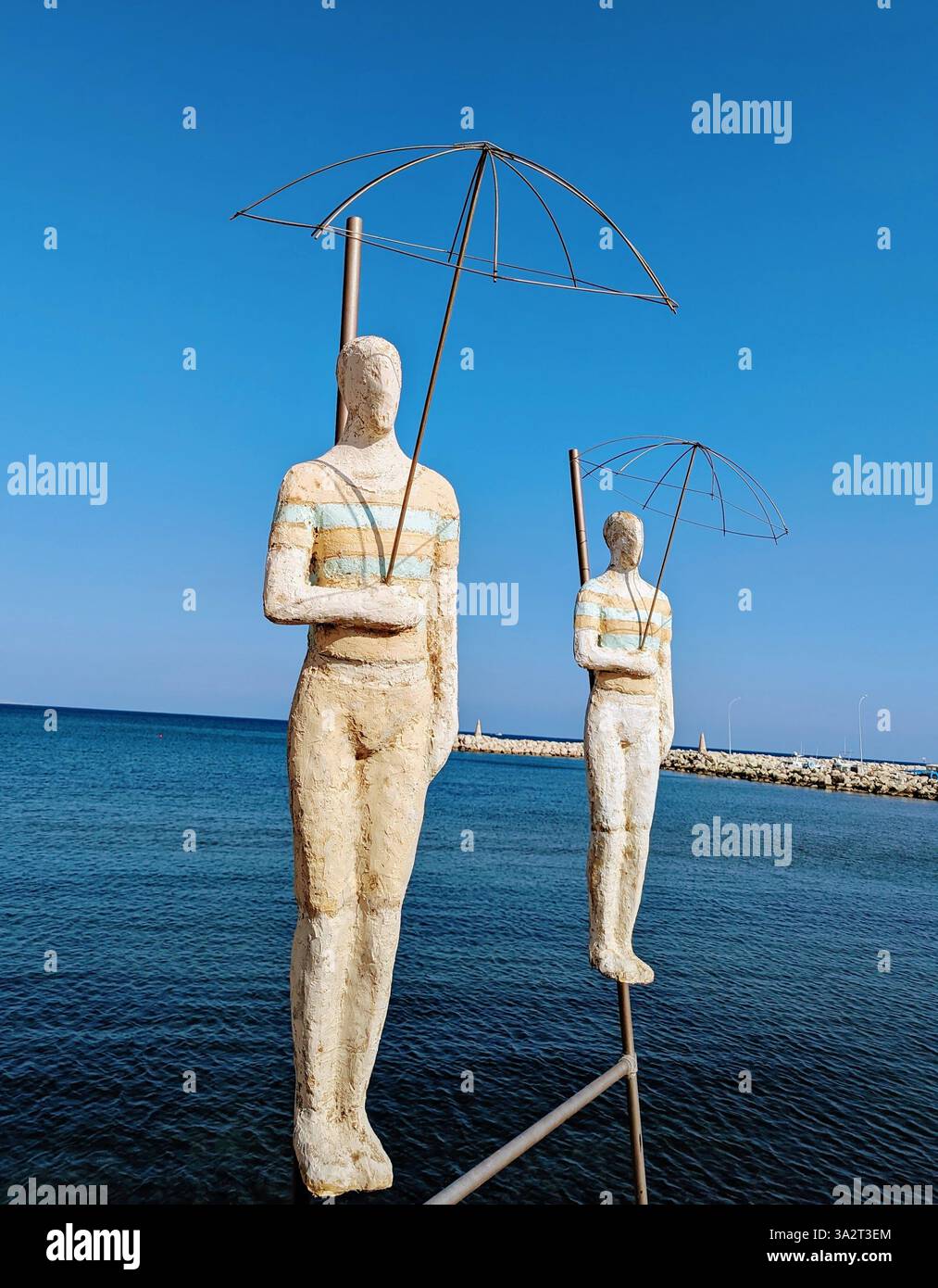 Am Kastella Beach stehen zwei künstlerische Statuen am Meer, die Regenschirme mit Drahtgestellen vor dem blauen Himmel halten und Einsamkeit und Reflexion symbolisieren. Stockfoto