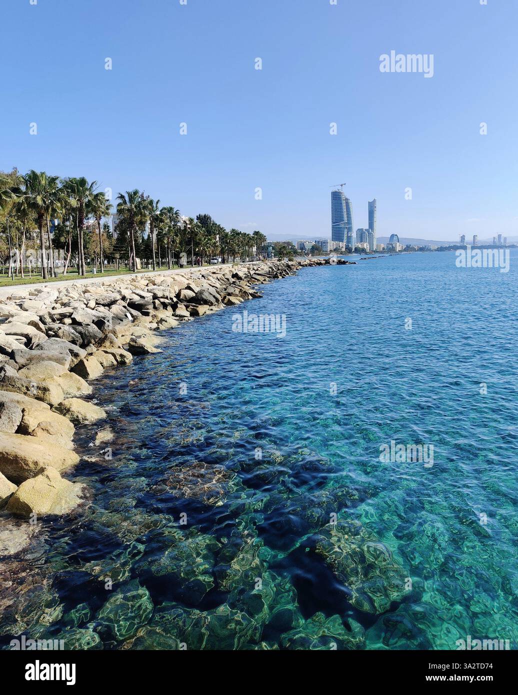 Blick entlang der Limassol Promenade in Zypern, mit klarem türkisfarbenem Wasser, Palmen und modernen Hochhäusern am Horizont. Stockfoto
