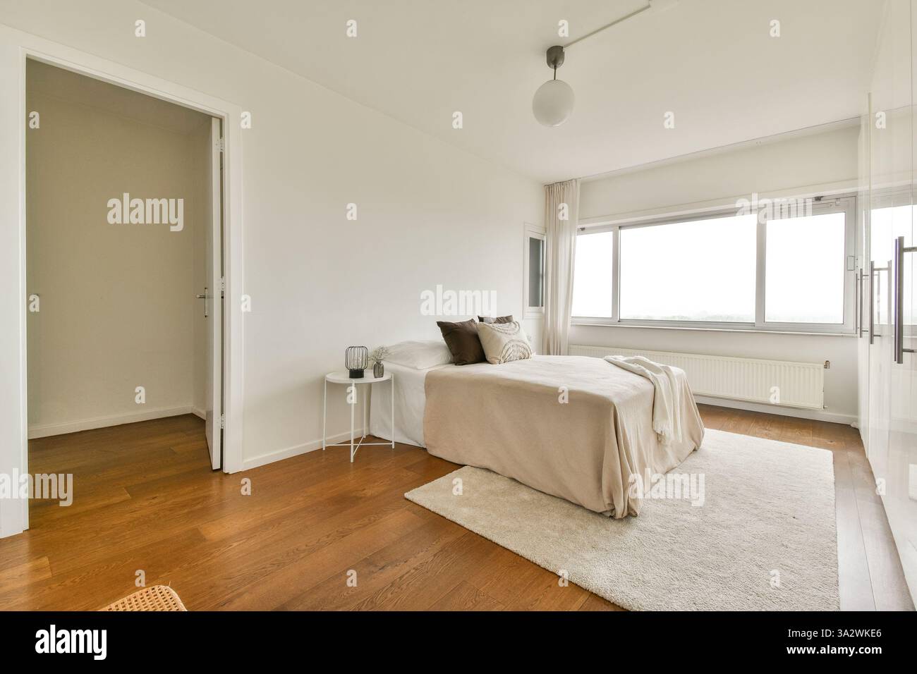 Ein wunderschön gestaltetes, minimalistisches Schlafzimmer mit neutralen Farben, Holzfußboden und großen Fenstern, die natürliches Licht einladen, schaffen eine ruhige Atmosphäre. Stockfoto