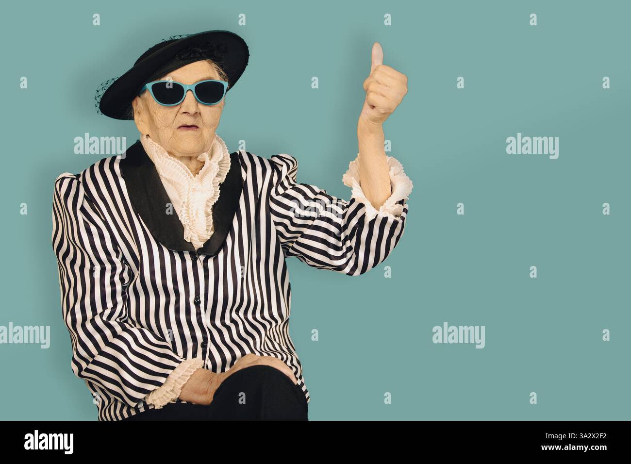 Eine ältere Frau in einem modischen Streifenoutfit und einer Sonnenbrille zeigt sich vor einem schlichten Hintergrund mit dem Daumen nach oben und verkörpert Selbstvertrauen und Sinn Stockfoto