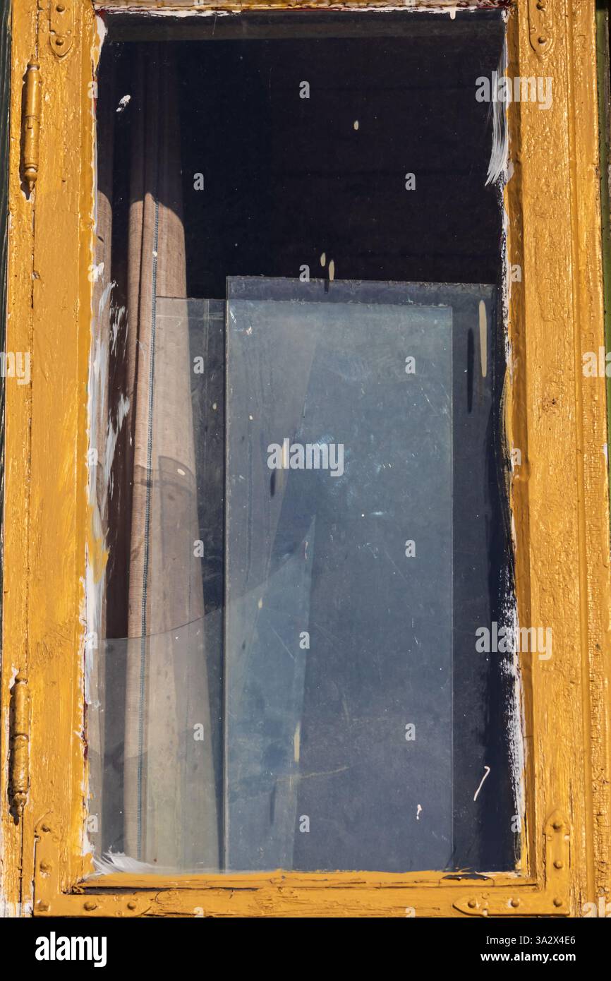 Ein gelber hölzerner Fensterrahmen mit Pinselspuren, der schmutziges, gestapeltes Glas dahinter offenbart, das ein vergessenes, verlassenes Gefühl weckt Stockfoto Ein gelber hölzerner Fensterrahmen mit Pinselspuren, der schmutziges, gestapeltes Glas dahinter offenbart, das ein vergessenes, verlassenes Gefühl weckt Stockfoto