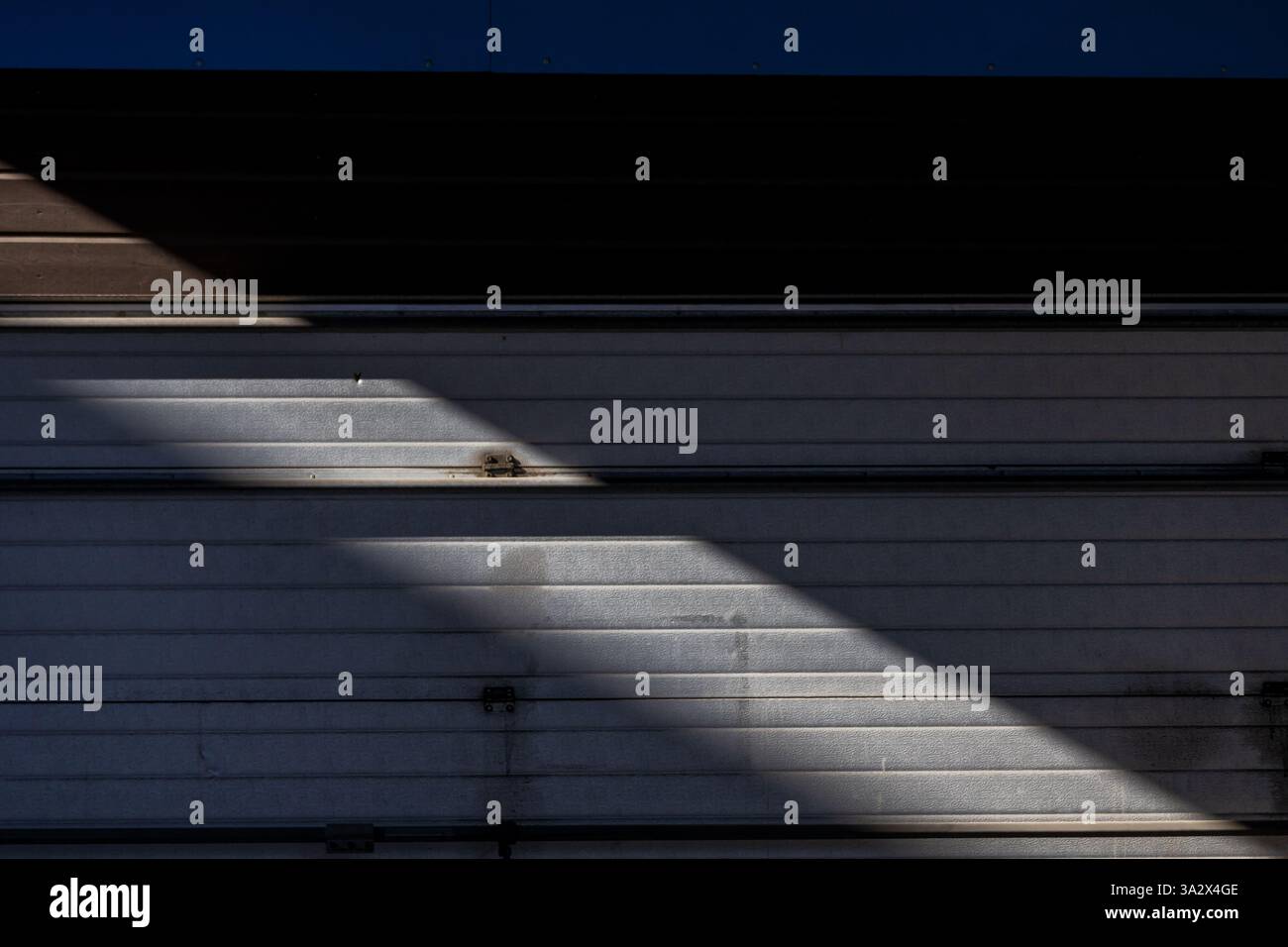Ein aufrollbares Garagentor befindet sich meist im Schatten, unterbrochen von einem einzigen diagonalen Lichtstrahl, der subtile Texturen freigibt und dramatischen Kontrast verleiht Stockfoto