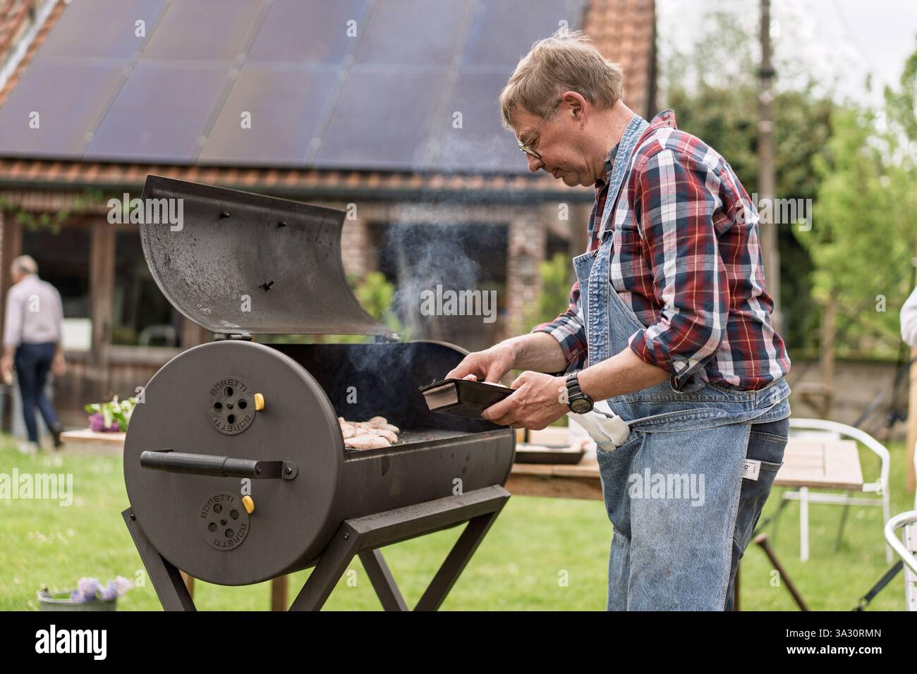 Senior Mann kocht auf einem Gartengrill, genießt Outdoor-Kochen und Freizeitaktivitäten. Reifer Mann im Overall, der im Garten Essen grillt, aktiv Stockfoto