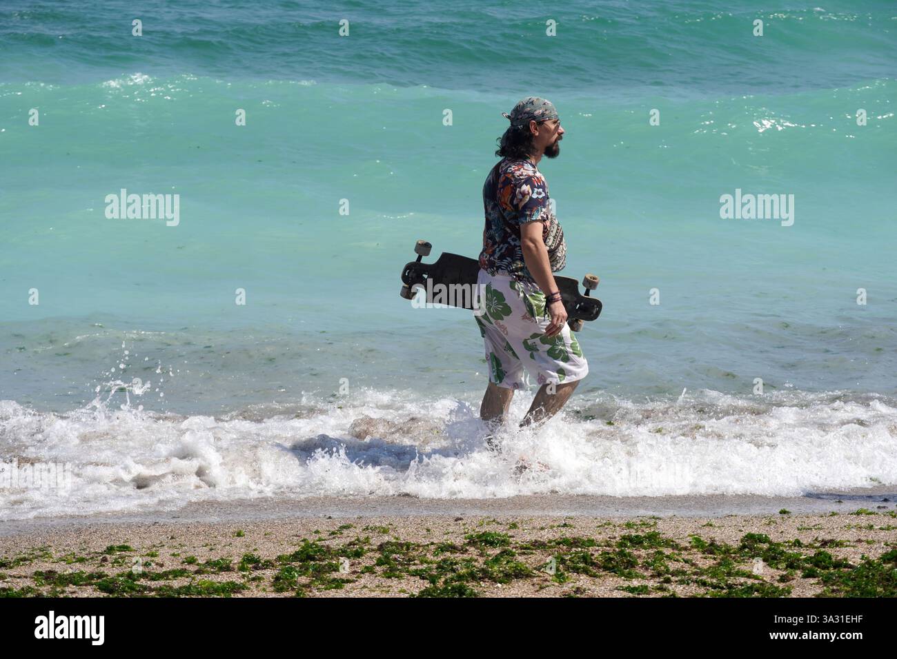 Vama Veche, Rumänien – 27. August 2019: Mann in tropischem Sommeroutfit, mit Skateboard, spaziert am Strand durch schaumige Meereswellen Stockfoto