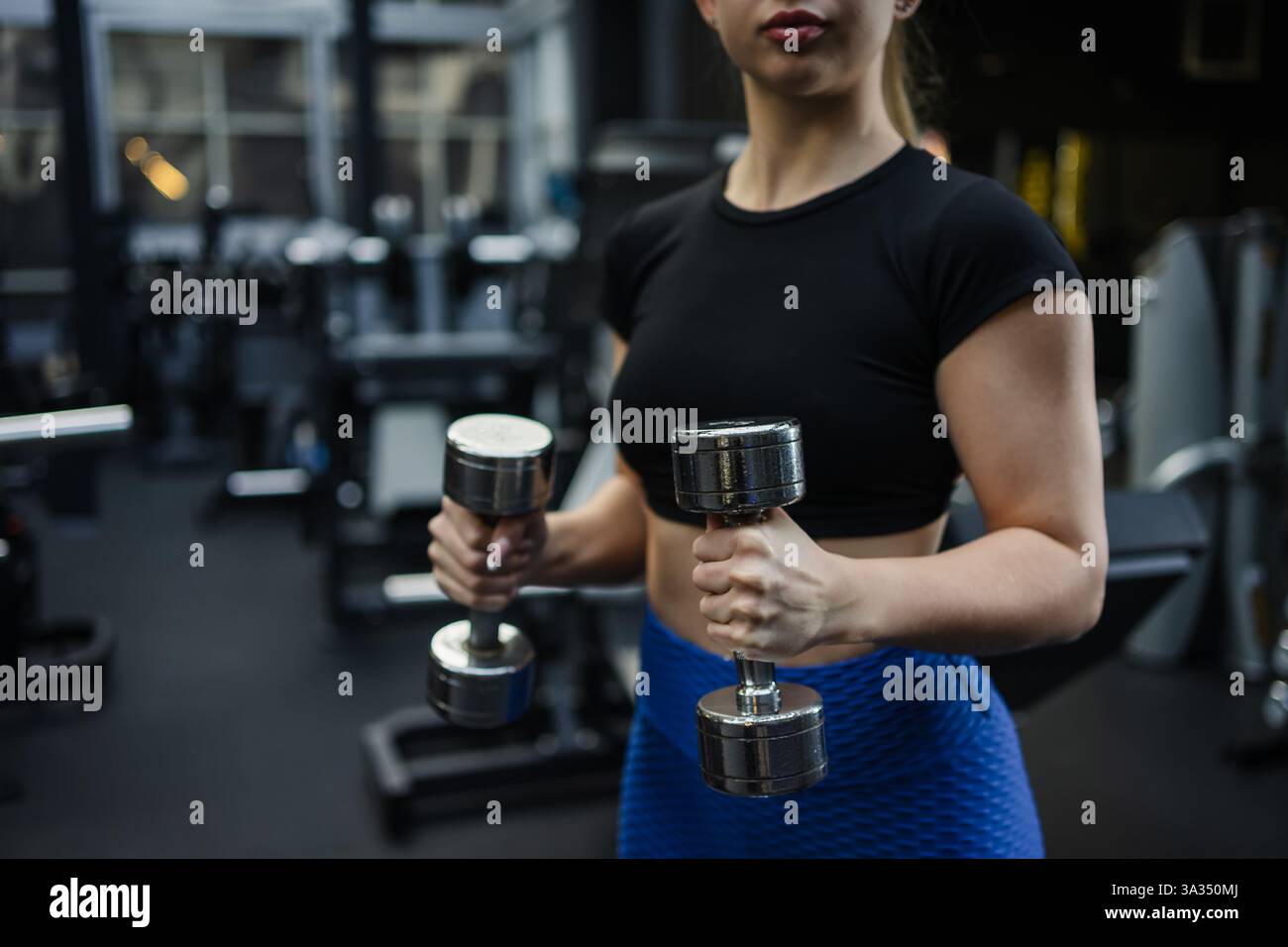 Eine blonde Frau in sportlicher Ausrüstung hebt Kurzhanteln in einem geräumigen, modernen Fitnessstudio mit professioneller Ausrüstung. Das Bild erfasst Stärke und Abschreckung Stockfoto