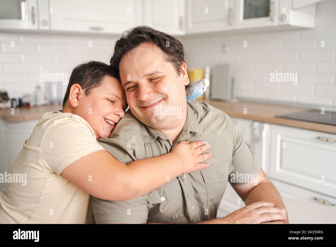 Eine herzerwärmende Szene, in der ein Vater und ein Sohn in einer hellen Küche eine fröhliche Umarmung teilen. Ein kleiner Vogel sitzt auf der Vaterschulter und fügt das c hinzu Stockfoto