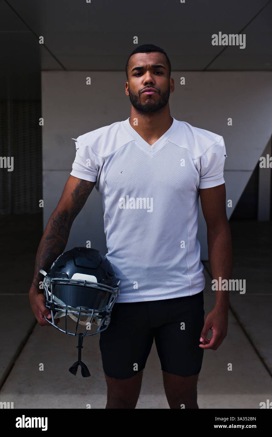 Ein fokussierter American Football-Spieler steht draußen und hält seinen Helm in einem weißen Trikot. Sein entschlossener Ausdruck verkörpert den Geist des Sp Stockfoto