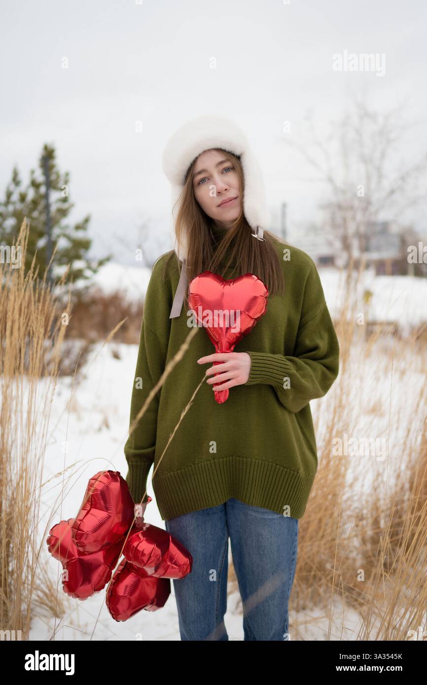 Frau in einem grünen Pullover, die rote Herzballons in einem verschneiten Park hält, umgeben von trockenem Gras, die Wärme und Liebe zur Kälte verkörpern. Stockfoto