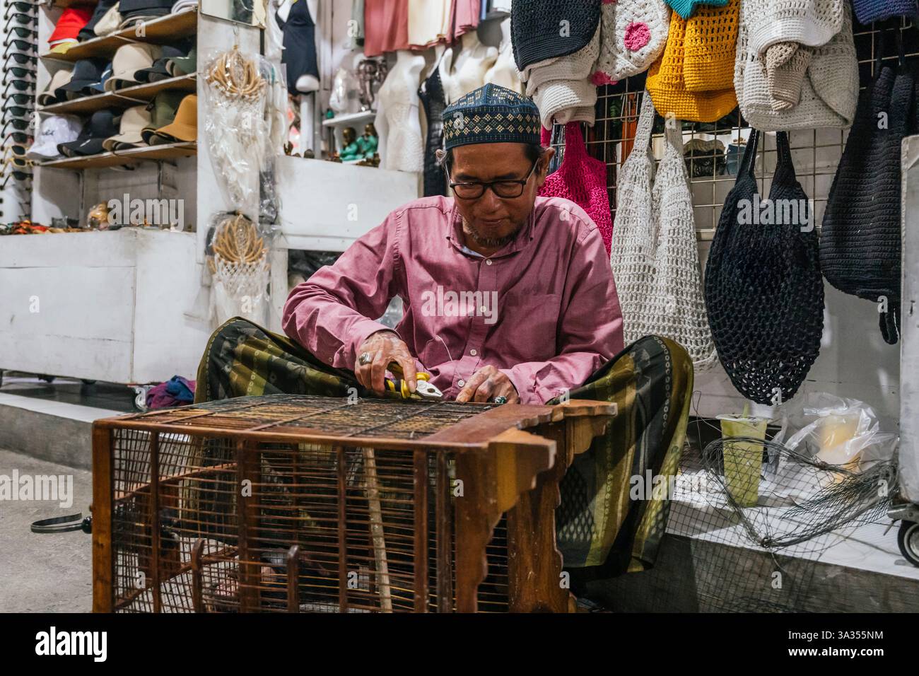 Ein balinesischer Handwerker, der ein Produkt geschickt herstellt, umgeben von bunten handgemachten Häkeltaschen in einer lebhaften Marktumgebung. Die Szene spiegelt trad wider Stockfoto
