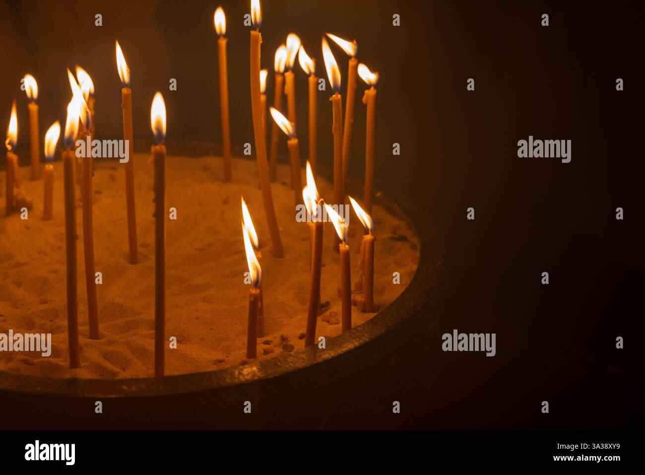 Die Kerzen sind in Sand angeordnet und ihre Flammen leuchten warm vor einem dunklen kirchlichen Hintergrund, was Gefühle von Spiritualität, Gelassenheit und Reflexion hervorruft Stockfoto