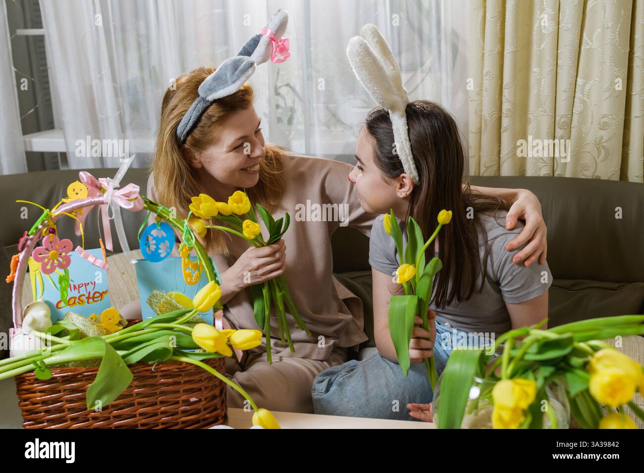 Mutter umarmt ihre Tochter. Eine Mutter und Tochter mit Hasenohren teilen einen freudigen Moment, während sie gelbe Tulpen in einer gemütlichen Osterstimmung halten. Stockfoto