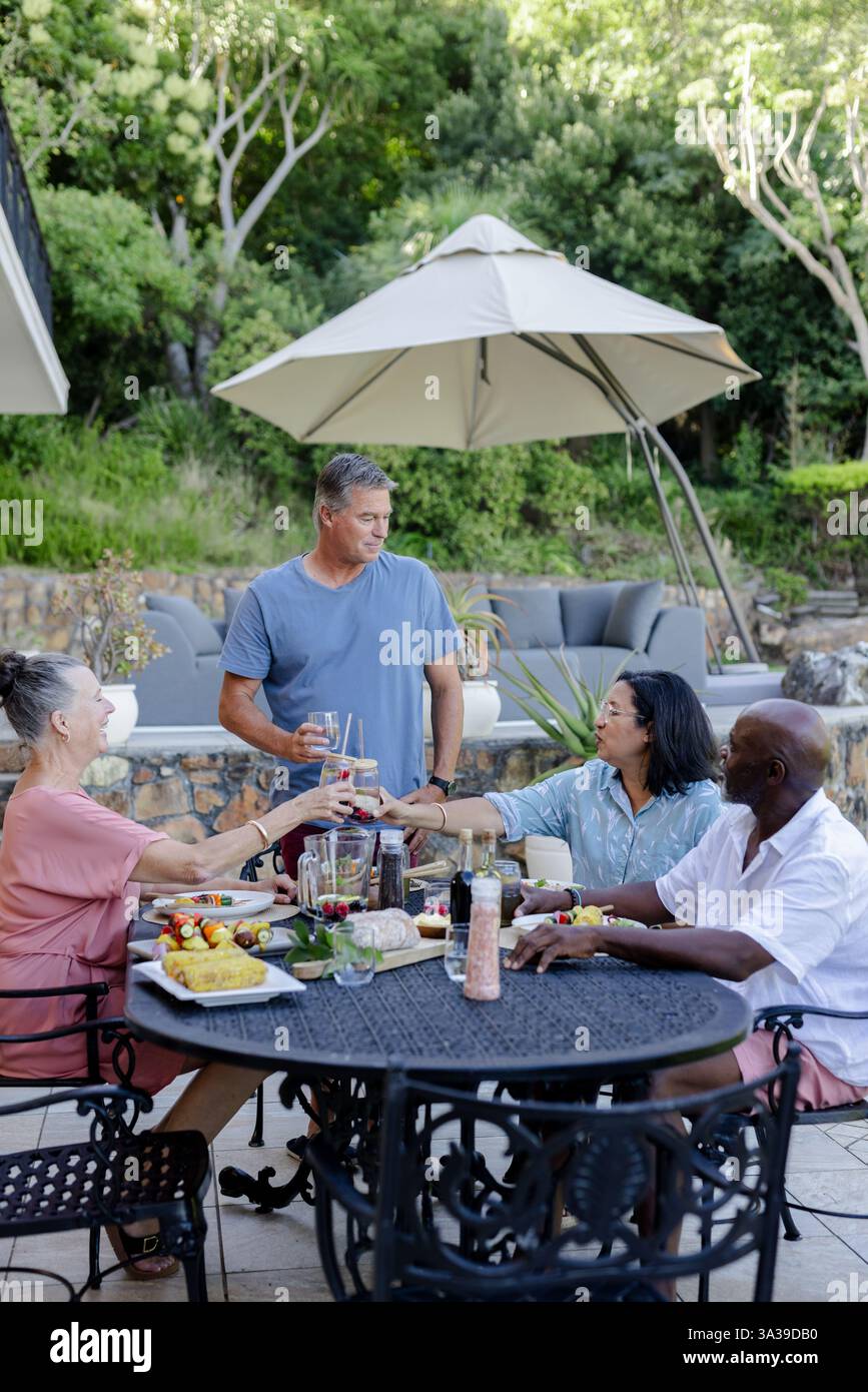 Eine Gruppe verschiedener älterer Freunde genießt ein Mittagessen im Freien, teilt Getränke und lacht im Garten Stockfoto