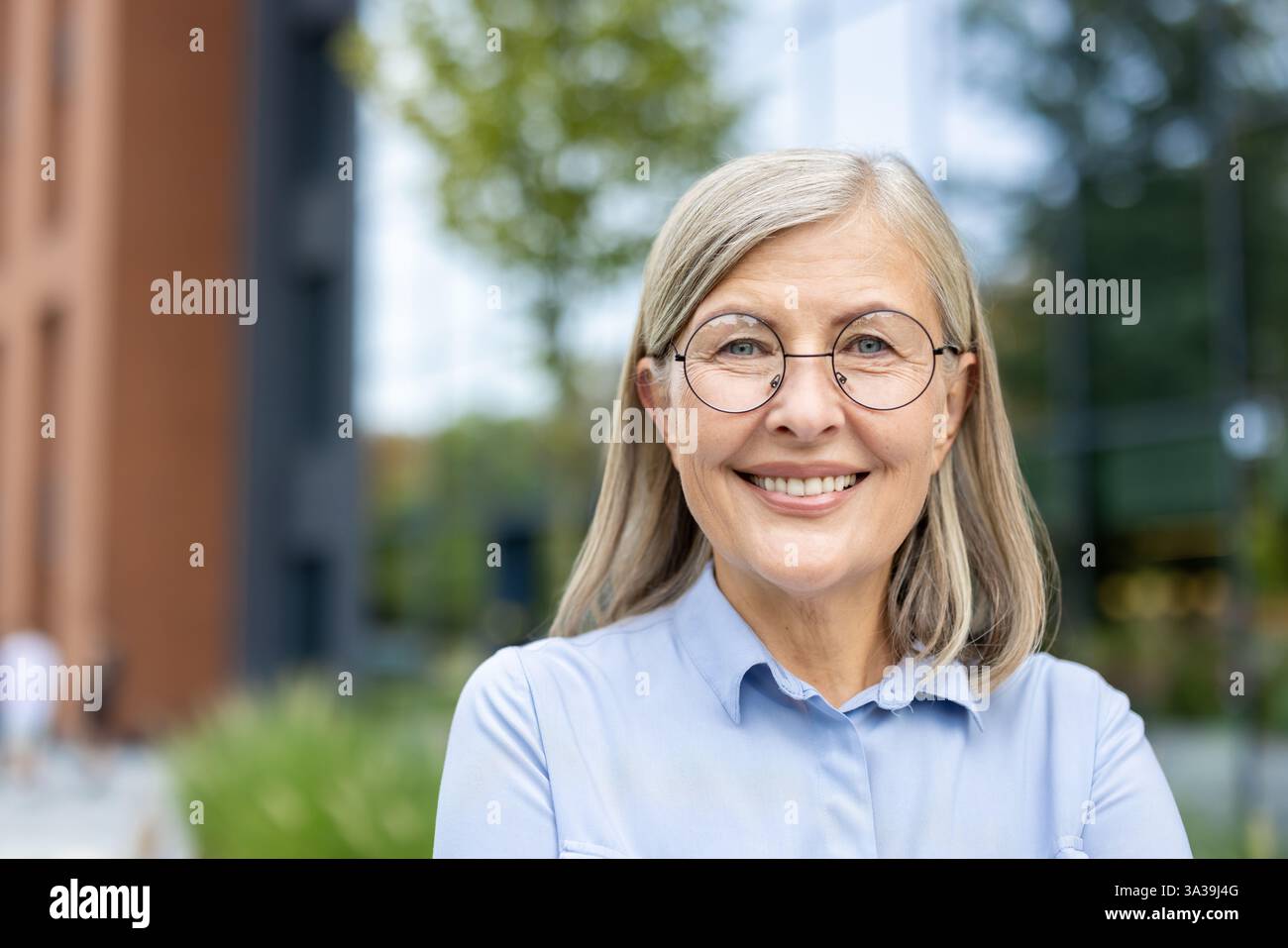 Ein Porträt einer Reifen, selbstbewussten Geschäftsfrau, die draußen lächelt, mit moderner Architektur und Grün im Hintergrund. Stockfoto
