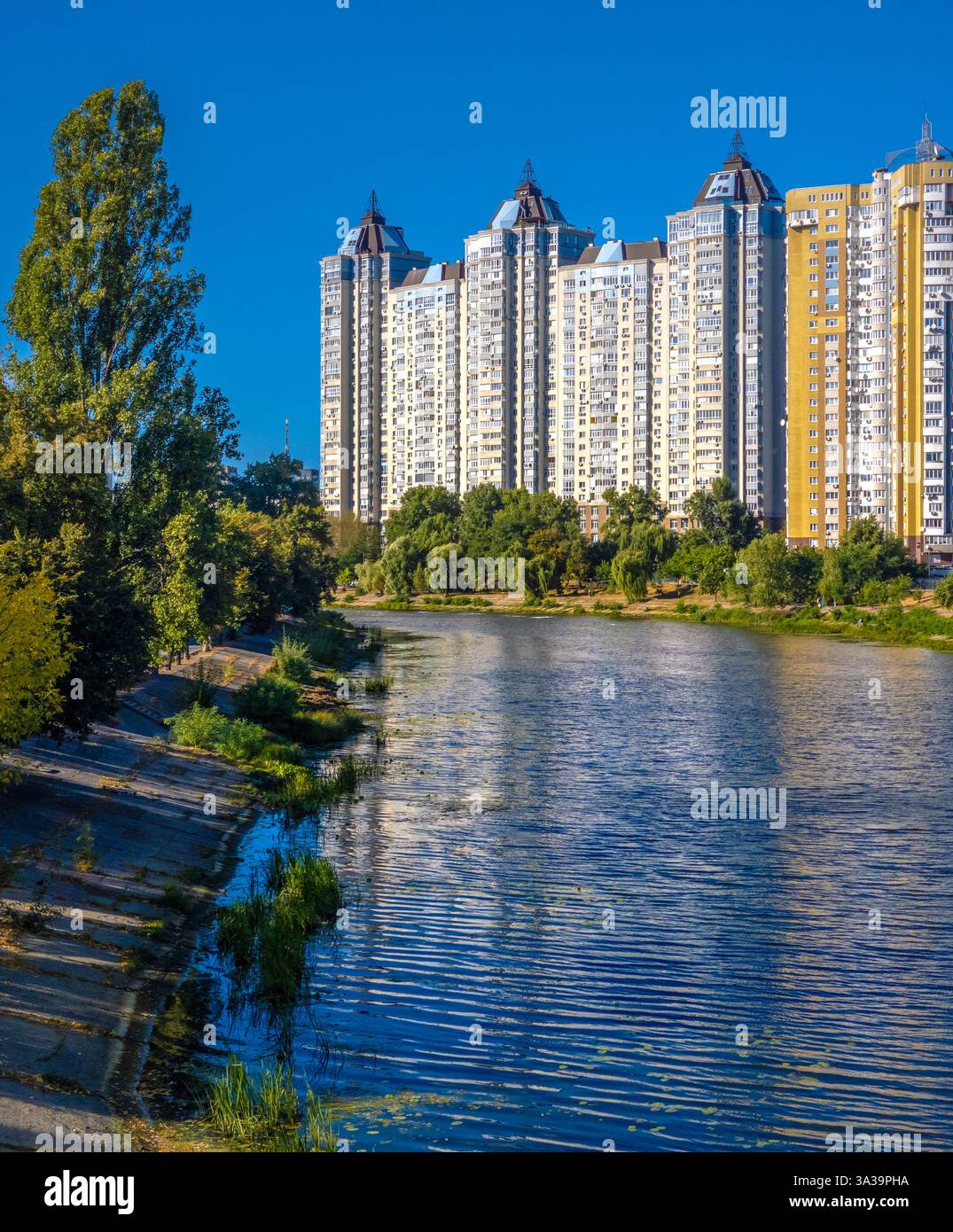 Wunderschöne Gebäude erheben sich über einem ruhigen Fluss, mit üppigem Grün am Ufer an einem sonnigen Tag, was einen friedlichen städtischen Zufluchtsort schafft. Stockfoto