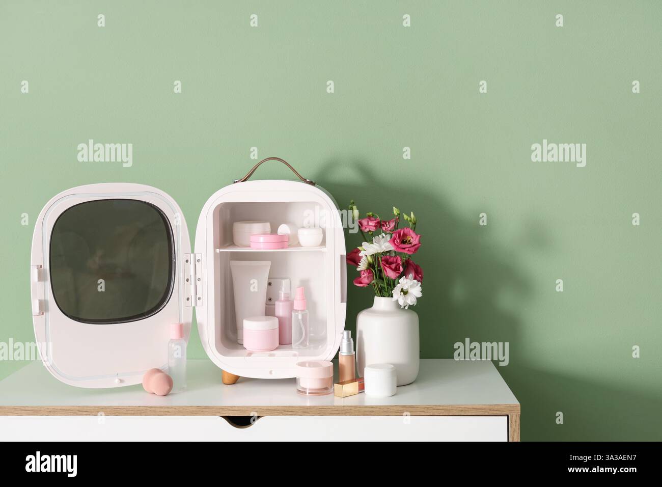 Öffnen Sie den kleinen Kühlschrank mit Kosmetikprodukten und Blumen in der Vase auf der Kommode vor grünem Hintergrund. Nahaufnahme Stockfoto