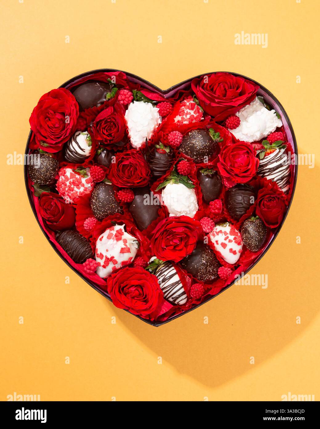 Luxuriöse Box in Herzform mit roten Rosen, Schokoladenerdbeeren und Himbeeren. Ein atemberaubendes Geschenk-Arrangement Stockfoto