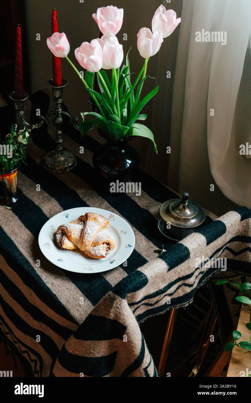 Ein Tisch ist mit einem Teller mit Gebäck, rosa Tulpen und Kerzen gedeckt, was eine ruhige Atmosphäre schafft, die perfekt für einen gemütlichen Nachmittagstee ist. Stockfoto