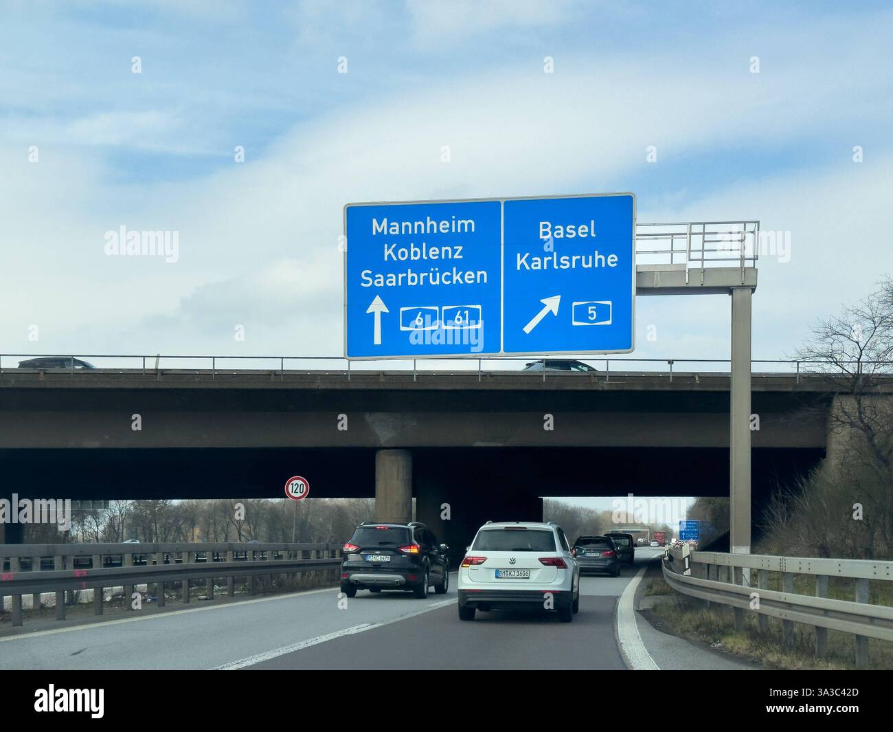 Deutschland, Europa: Autobahnszene mit Wegweiser Verkehr auf einer deutschen Autobahn mit Hinweisschildern in Richtung Mannheim, Koblenz, Saarbrücken A6/A61 und Basel, Karlsruhe A5. Fahrzeuge fahren unter einer Brücke hindurch, die Fahrbahn ist gut frequentiert. Geschwindigkeit ist auf 120 km/h begrenzt. Typische Verkehrssituation auf einer deutschen Autobahn. *** Deutschland, Europa Autobahnszene mit Beschilderung Verkehr auf einer deutschen Autobahn mit Beschilderung in Richtung Mannheim, Koblenz, Saarbrücken A6 A61 und Basel, Karlsruhe A5 Fahrzeuge fahren unter einer Brücke, die Straße ist voll Geschwindigkeit ist begrenzt auf Stockfoto