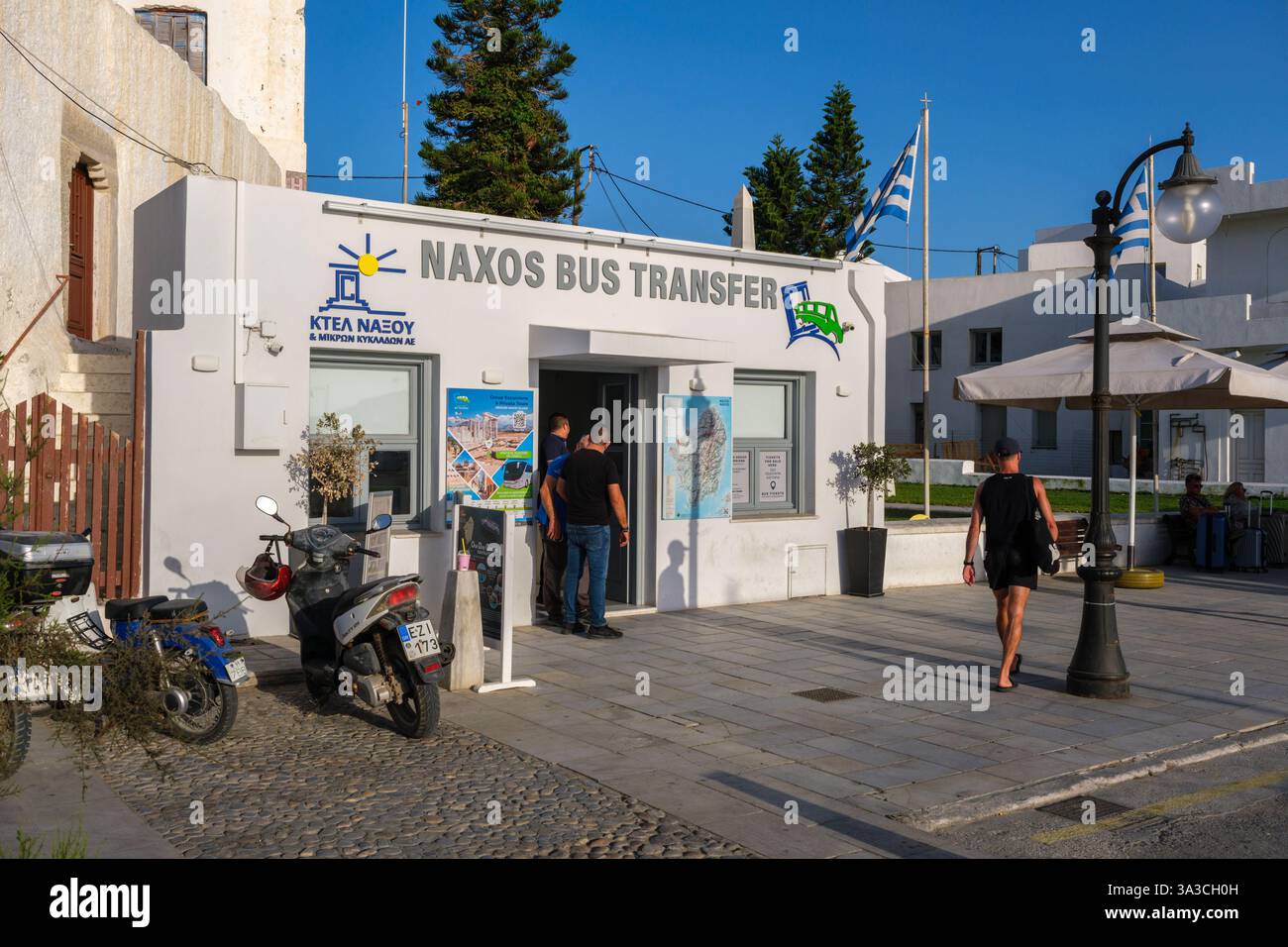 Naxos, Griechenland - 4. Oktober 2024: Naxos Bus Transfer Office in Naxos Chora, betrieben von KTEL Naxos, mit Personen und Motorrollern in der Nähe unter einem klaren bl Stockfoto