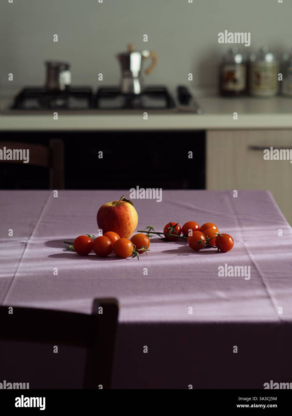 Kirschtomaten und ein Apfel auf einer lila Tischdecke, vertikaler Weitwinkelblick auf eine gemütliche italienische Küche Stockfoto