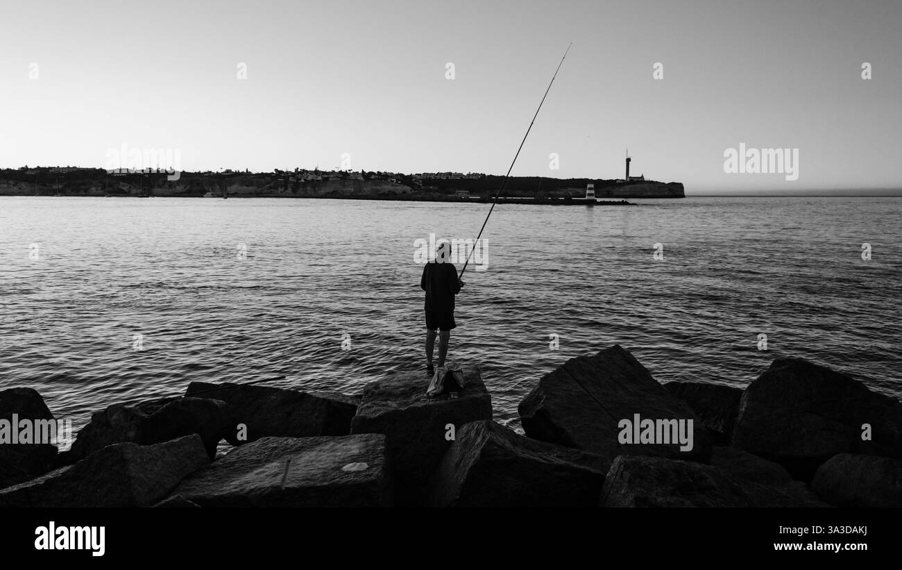 Zeitlose Schwarzweiß-Angelszene: Ein einsamer Angler wirft seine Linie von zerklüfteten Felsen vor einer ruhigen Küstenkulisse. Stockfoto