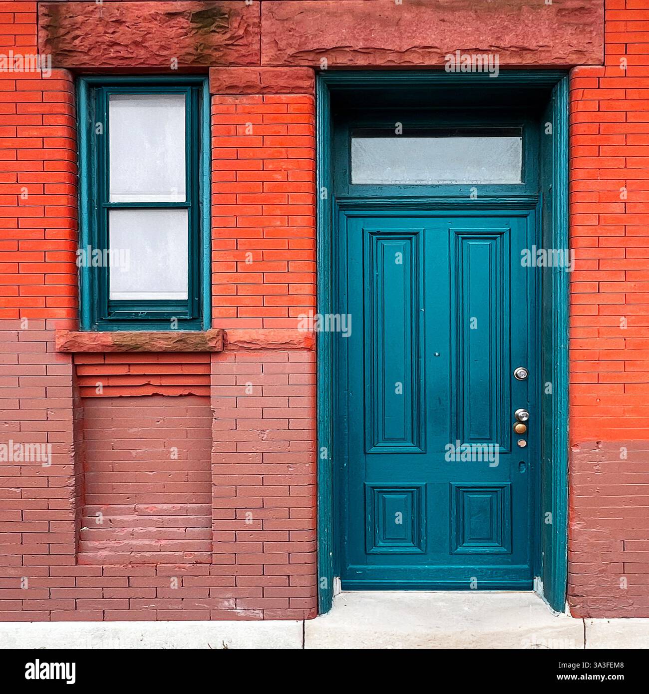 Teal-Holztür mit einem Heckfenster in einer historischen rot-braunen Ziegelfassade mit kontrastierenden Texturen und architektonischen Details. - Smartphone-aufgenommenes Stockfoto