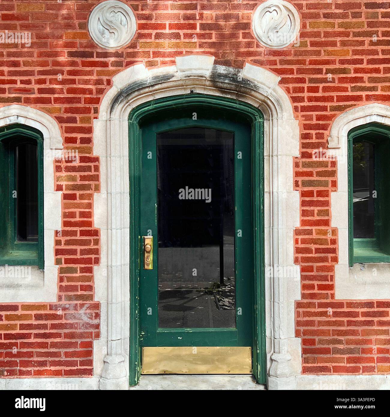 Grüne Tür im gotischen Stil, die sich in einer verzierten roten Ziegelstein- und Kalksteinfassade befindet, mit Bogenfenstern und dekorativen Steinmedaillons. - Smartphone-aufgenommenes Stockfoto