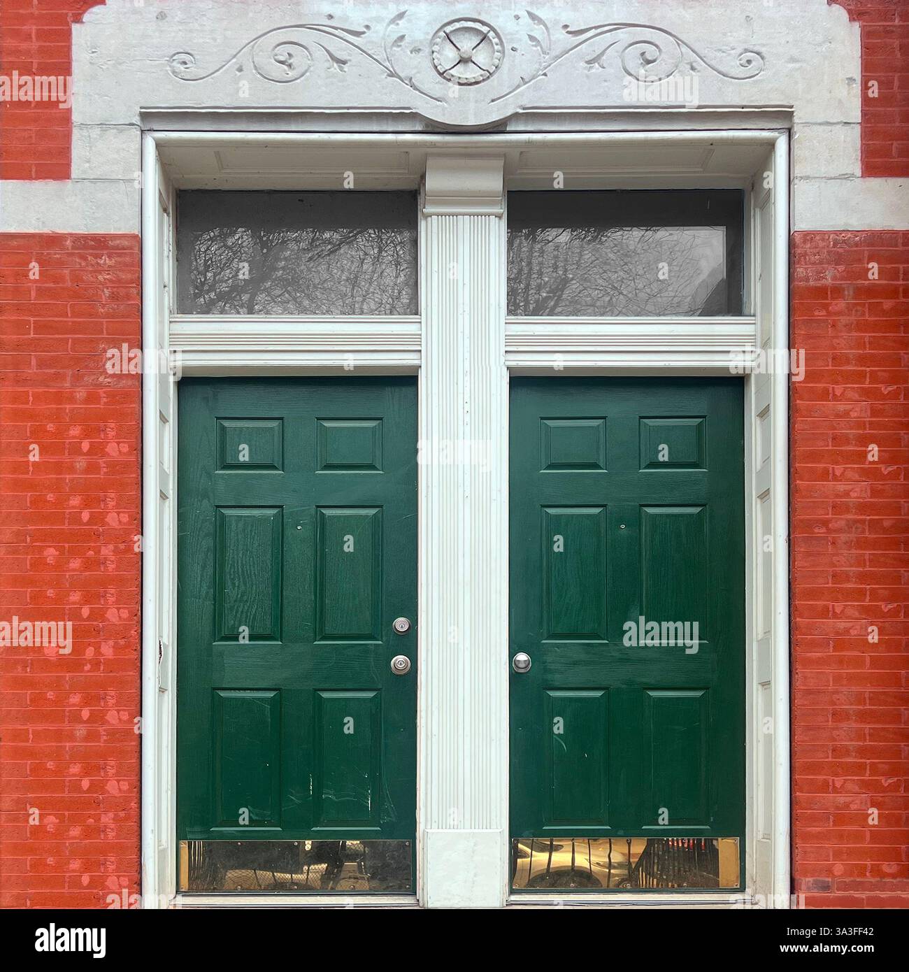 Grüne Doppeltüren mit klassischem Paneeldesign, eingerahmt von weißen Pilastern und Heckfenstern, die vor einer roten Ziegelfassade mit dekorativem Mauerwerk stehen. - Smartphone-aufgenommenes Stockfoto