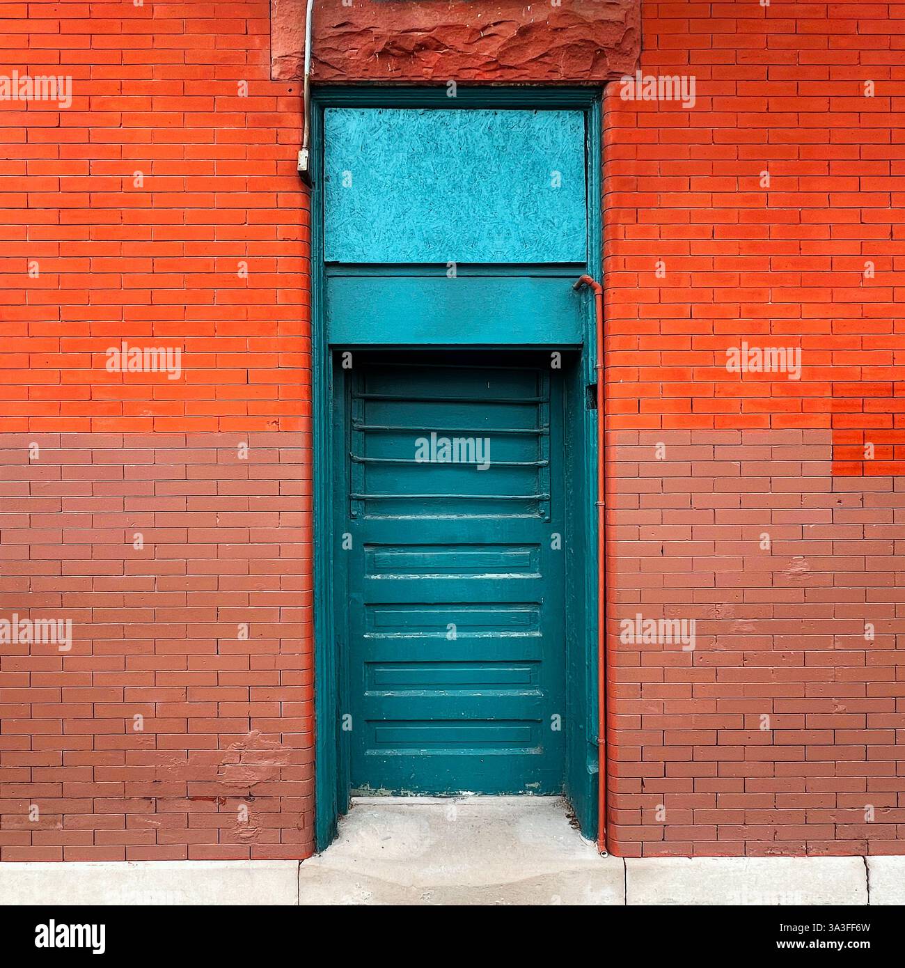 Eine verwitterte, türkisfarbene Tür in einer kräftigen rot-braunen Ziegelwand, die einen auffälligen Kontrast von Farben und Texturen schafft. - Smartphone-aufgenommenes Stockfoto