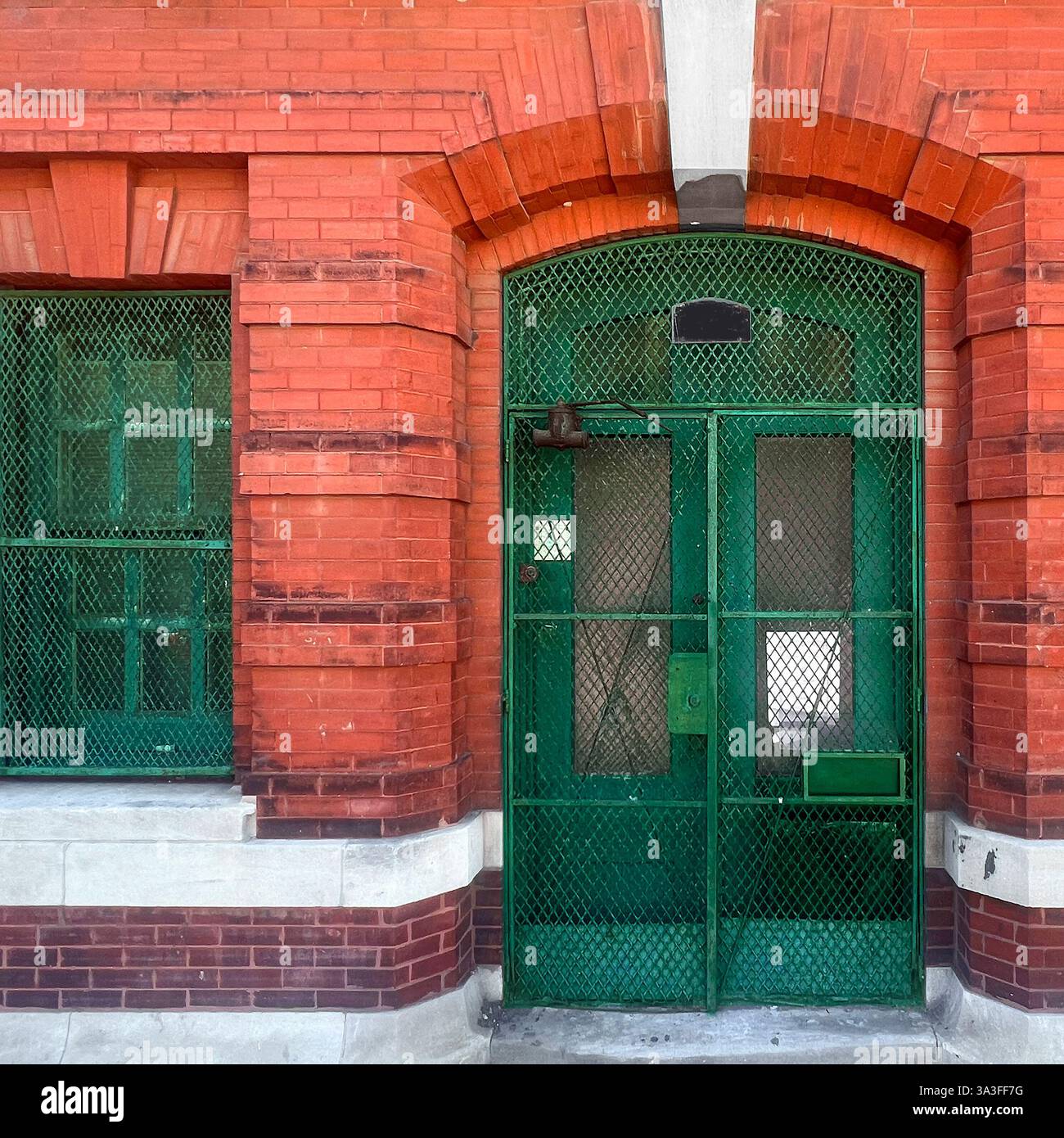 Ein altes rotes Backsteingebäude mit einem bogenförmigen Eingang mit einem verwitterten grünen Metall-Sicherheitstor und Fenstergittern, die an eine industrielle Ära erinnern. Stockfoto