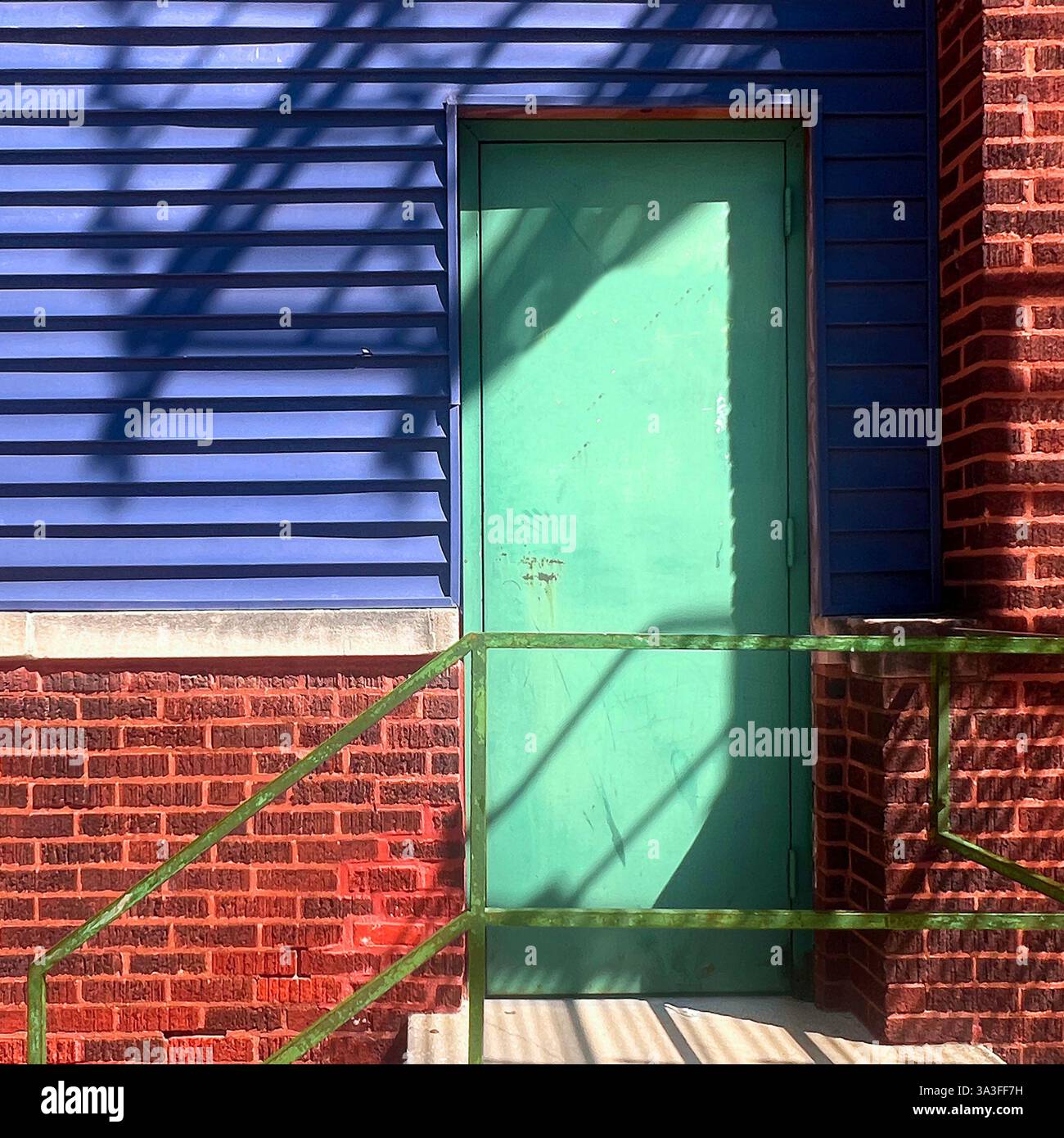 Eine verwitterte grüne Metalltür in einem Industriebau mit blauem Abstellgleis, rotem Backstein und grünem Geländer wirft dramatische Schatten in die Sonne. - Smartphone-aufgenommenes Stockfoto