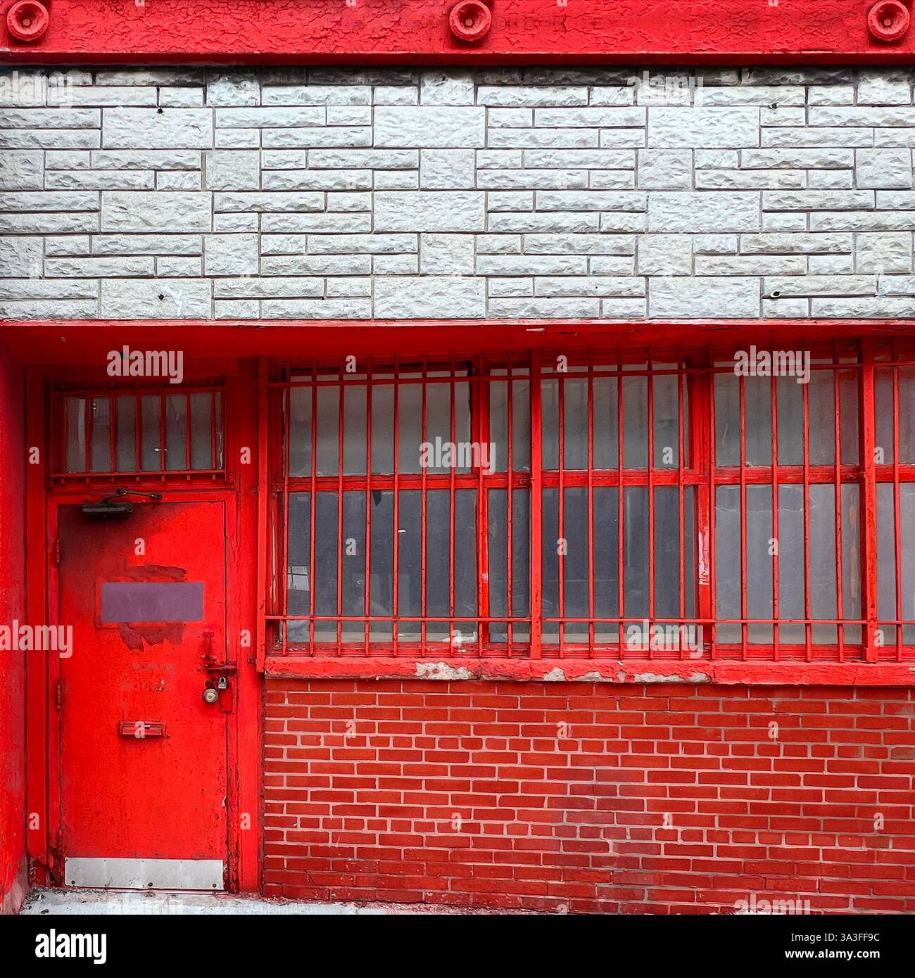 Eine verwitterte rote Metalltür mit vergitterten Fenstern in einem städtischen Backsteingebäude, die einen starken Kontrast zur weißen Steinfassade darüber schafft. Stockfoto