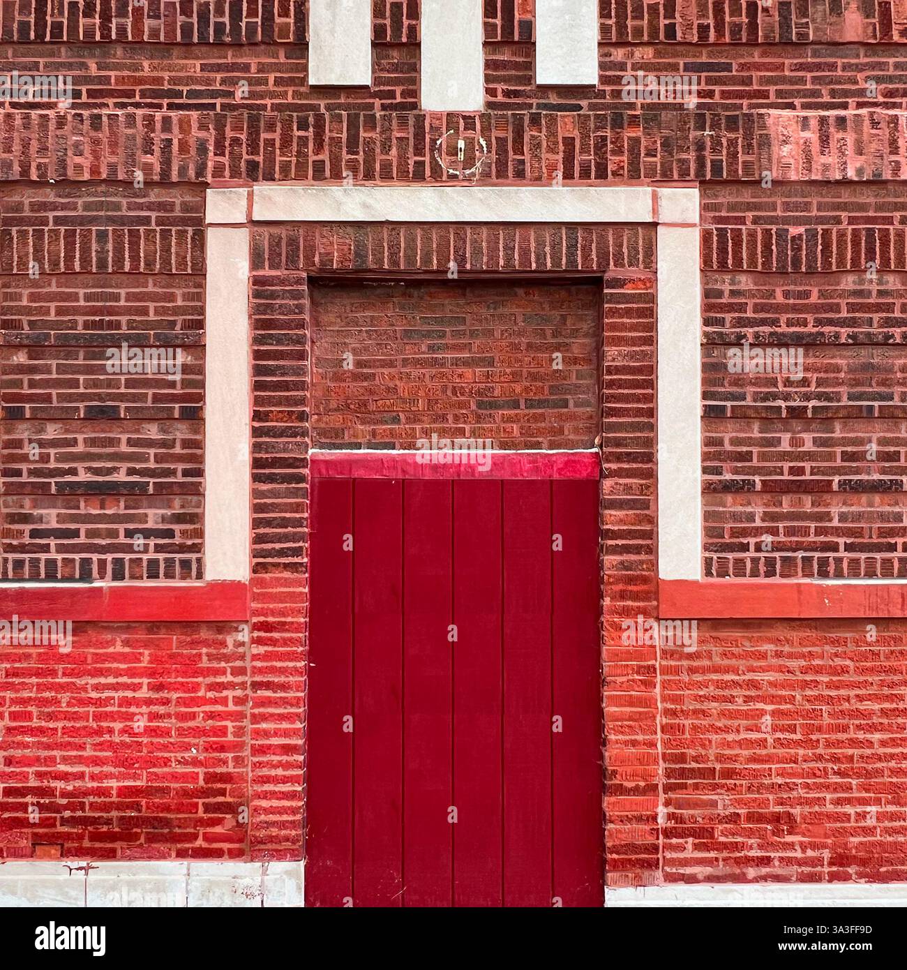 Industrieller Eingang aus rotem Holz innerhalb einer reich strukturierten roten Backsteinmauer, eingerahmt von kontrastierenden weißen Steinakzenten. - Smartphone-aufgenommenes Stockfoto
