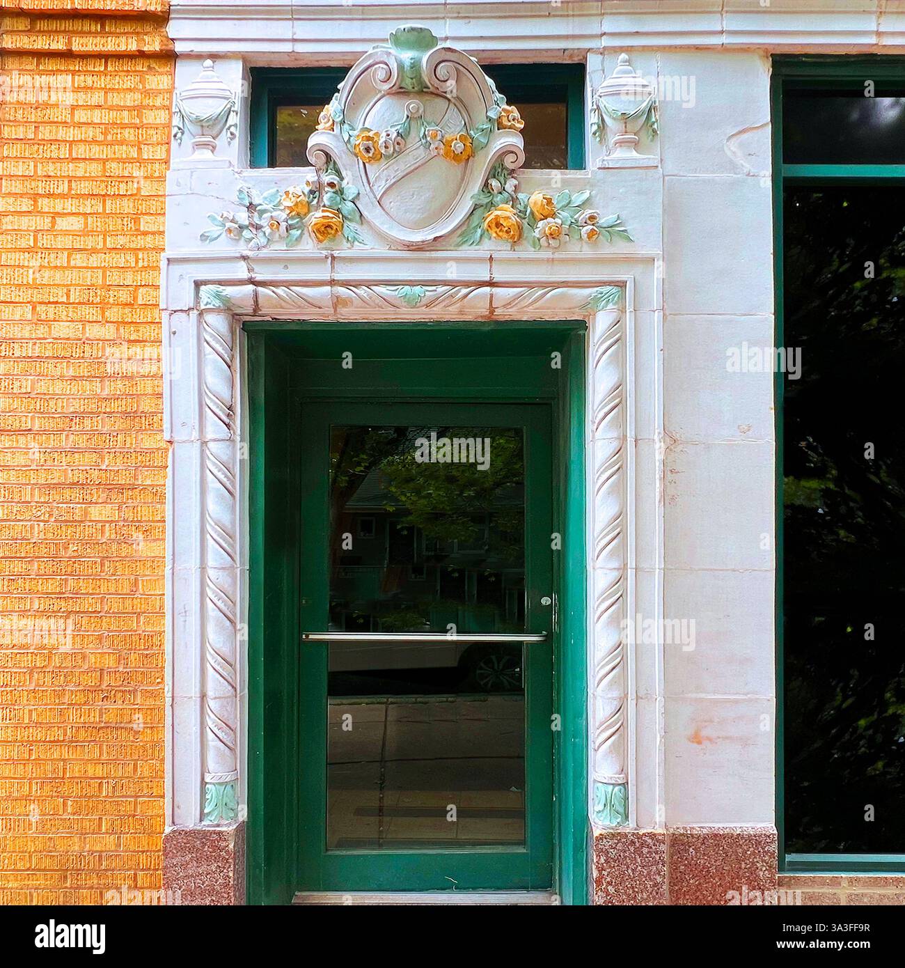 Ein historischer Gebäudeeingang mit verglasten Terrakotta-Details, mit einem dekorativen Wappen mit floralen Verzierungen über einer grünen Glastür. Stockfoto
